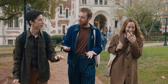 Three young people walking on a college campus, two men talking while a woman laughs covering her mouth, Gothic buildings behind.