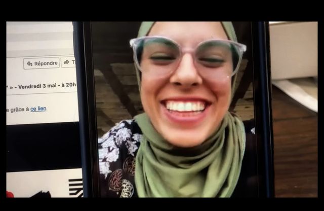 Smiling woman wearing an olive green hijab and clear-framed glasses, displayed on a phone screen next to a French email.