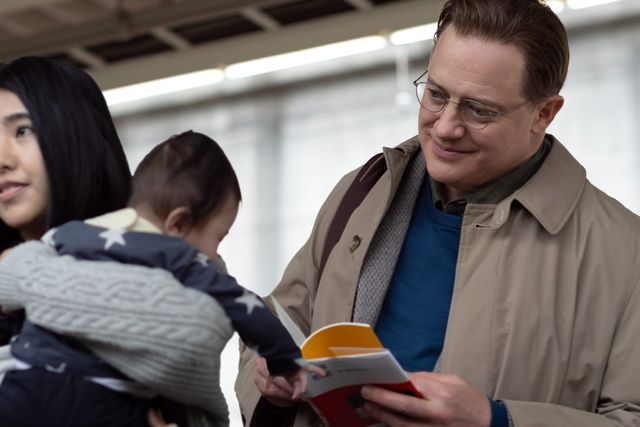 Man in glasses and tan coat reading a book smiles near woman holding a baby in a star-patterned outfit, indoor setting.