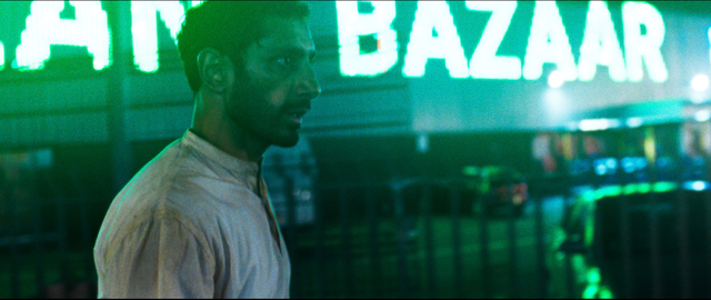 Man in white shirt seen in profile at night, illuminated by green neon "BAZAAR" sign glowing behind him.
