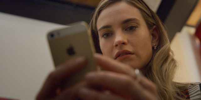 Young woman with blonde wavy hair looking at a gold iPhone with a focused, slightly concerned expression on her face.