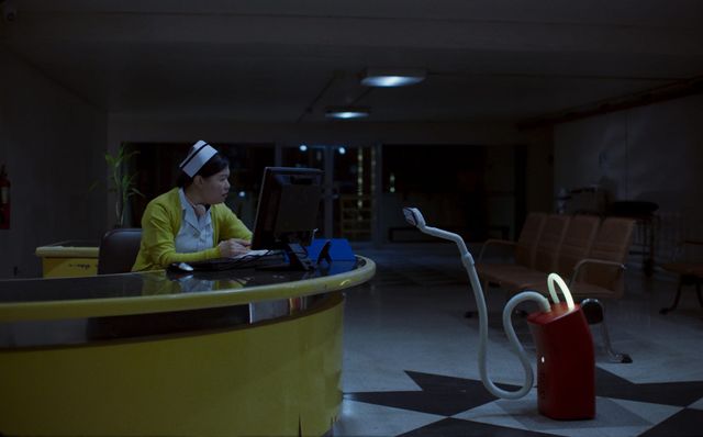 Nurse in yellow cardigan and white cap works at a dimly lit hospital reception desk at night, with a red robot nearby.