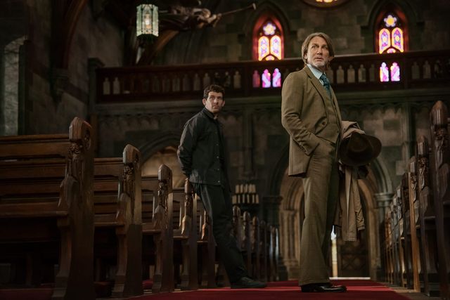 Two men standing in a Gothic church aisle between wooden pews; one in dark clothing, the other in a tan suit holding a hat.