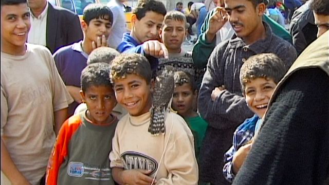 Group of smiling boys and teenagers crowded together outdoors on a street, some pointing at the camera, in a Middle Eastern setting.