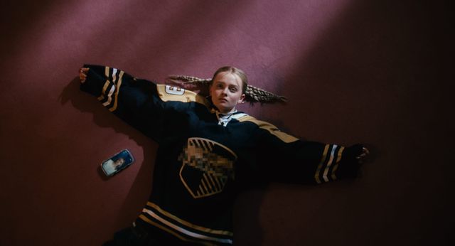 Young woman with braids lying on a dark red floor wearing an oversized black and gold hockey jersey, phone beside her.