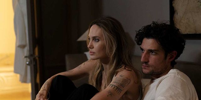Two people sitting close together in a dimly lit room; a tattooed woman with long hair and a man with dark curly hair.