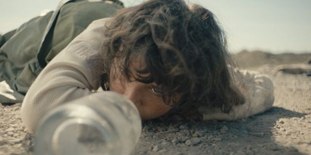 Person lying face-down on dry, dusty ground reaching for an empty water bottle, appearing exhausted and dehydrated in a desert setting.