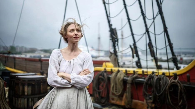 Woman in period colonial clothing with arms crossed standing on a sailing ship deck, wind blowing her hair, overcast sky.