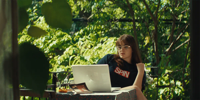 Young woman in glasses and dark "SPIN" t-shirt sits at outdoor table with laptop and magazine, surrounded by lush greenery.