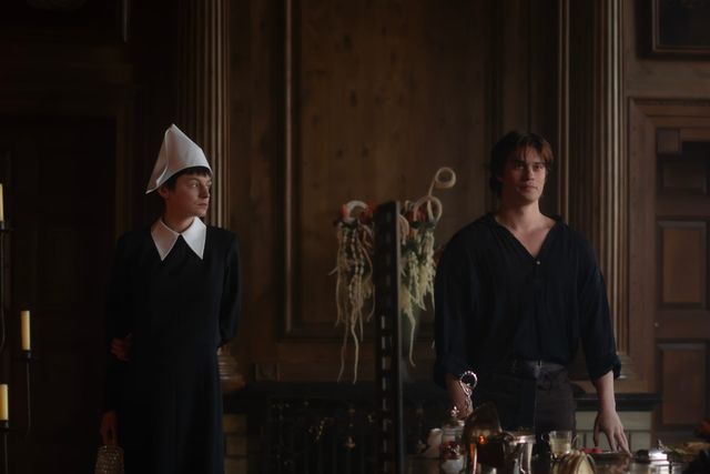 Two people in a dimly lit wood-paneled room; one wears a black dress with white collar and white pointed cap, the other a dark loose shirt.