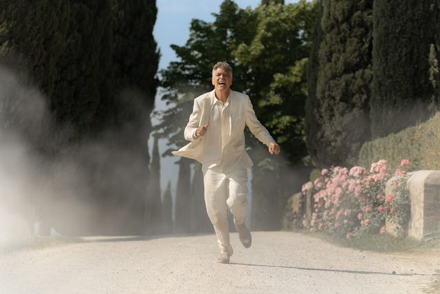 Man in white linen suit running urgently down a dusty cypress-lined path with pink roses and dust cloud behind him.