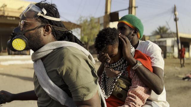 Three people on a street covering their faces from tear gas; one wears a gas mask, another a Rastafarian-colored beanie.