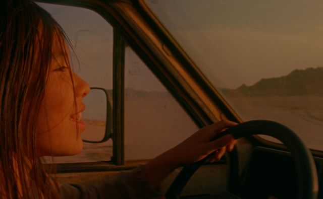 Young woman driving a vintage truck through a desert landscape at golden hour, eyes closed, hair blowing in the wind.