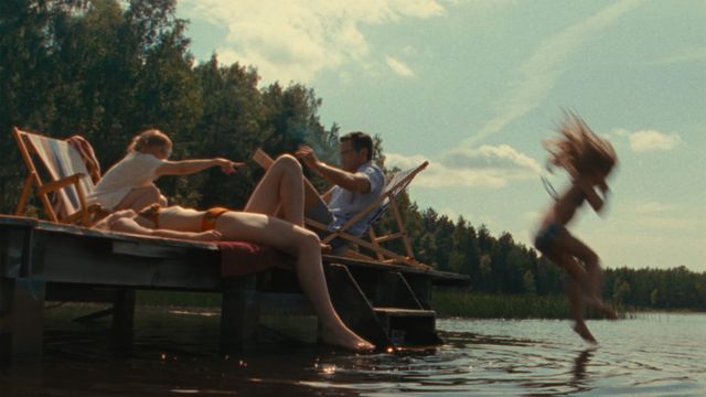 Family on a wooden lake dock in summer; adults relax in deck chairs while a child leaps into the calm water, hair flying.