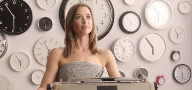 Woman wrapped in gray towel sitting at a typewriter against a wall covered with various clocks of different sizes and styles.