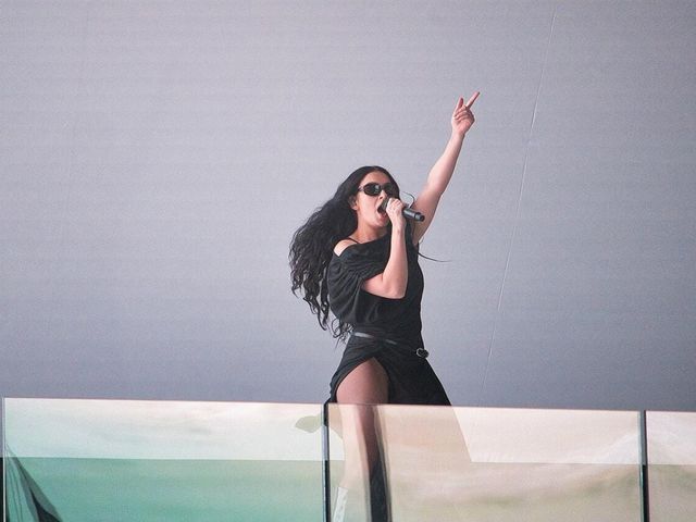 Woman with long dark curly hair and sunglasses singing into a microphone on stage, wearing black, pointing one finger up.
