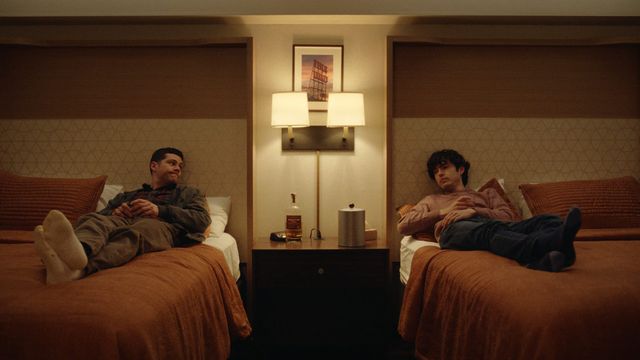 Two young men recline on separate twin beds in a warmly lit hotel room with a bottle of whiskey on the nightstand between them.