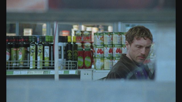 Man in dark jacket browsing a grocery store aisle with olive oil bottles, canned tomatoes, and boxed goods on shelves.