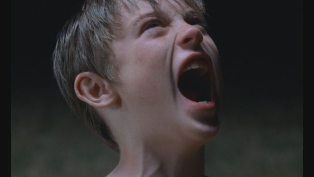 Close-up of a young boy with short blond hair screaming with his mouth wide open, head tilted back, against a dark background.