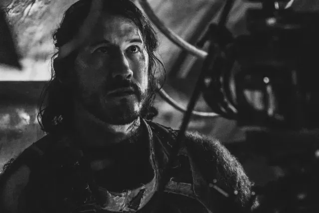 Black and white close-up of a man with long dark hair and a beard, wearing rugged clothing, looking upward intensely.