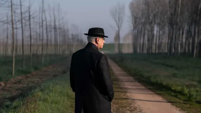 Elderly man in black overcoat and fedora stands on a rural dirt path lined with bare trees on a misty day, seen from behind.
