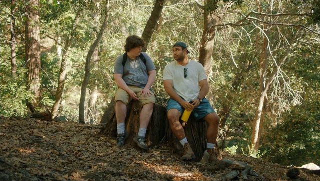 Two young men sitting on a tree stump in a wooded forest, one in a gray shirt and khaki shorts, the other in a white tee and blue shorts holding a yellow water bottle.