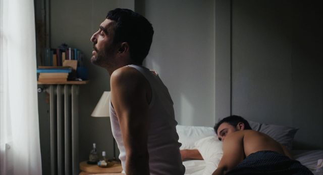 Man in white tank top sits on edge of bed looking pensive while another shirtless man sleeps face-down behind him.