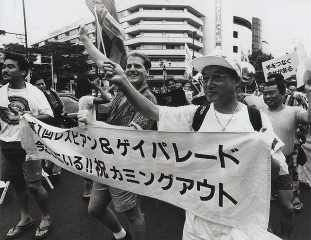 Black and white photo of marchers at a Japanese Lesbian & Gay Pride Parade holding a banner with Japanese text celebrating coming out.