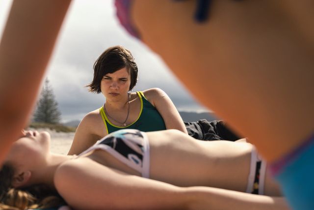 Young woman in green tank top sits on beach looking down at friend lying on sand in bikini, overcast sky behind them.