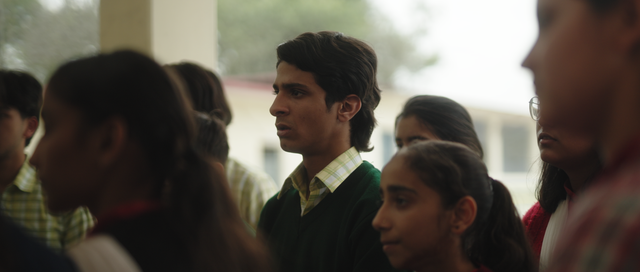 Young man in green sweater over plaid shirt stands among a crowd of students in what appears to be an Indian school setting.
