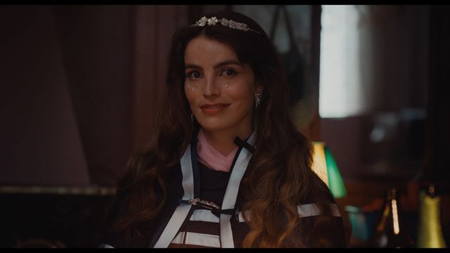 Young woman with long wavy brown hair, floral headband, teardrop face gems, dangling earrings, and striped outfit, smiling softly.