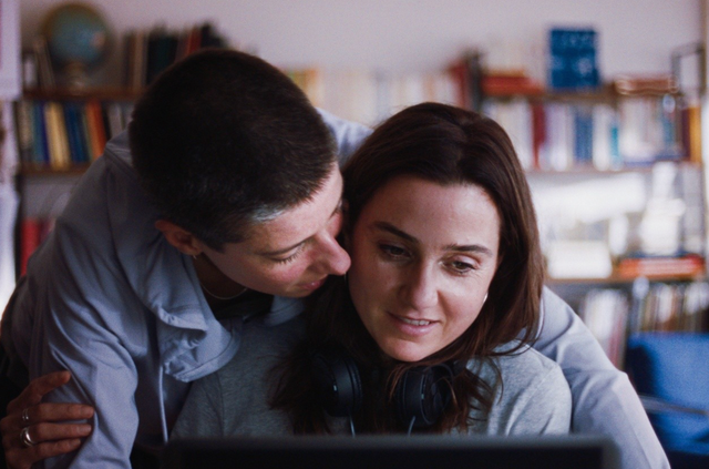 Two people looking at a laptop screen together, one leaning over the other's shoulder affectionately, bookshelves behind them.