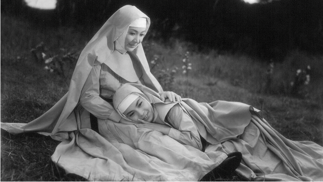 Two smiling women in nun habits sitting on grass, one reclining in the other's lap, in a black and white photograph.