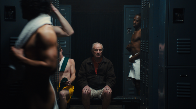 Older man sitting in a dimly lit locker room surrounded by younger athletes changing, with blue metal lockers behind them.