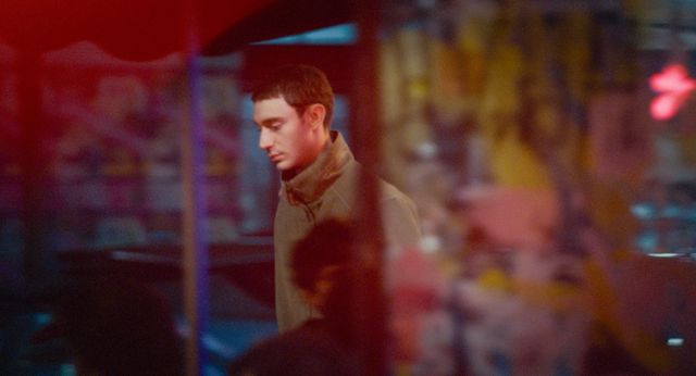 Young man in a tan jacket looking downward, photographed through glass with colorful red, blue, and yellow light bokeh.