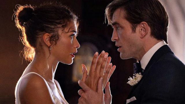 Two people face each other intimately, hands touching, she in a white dress with updo hair, he in a dark suit with boutonniere.