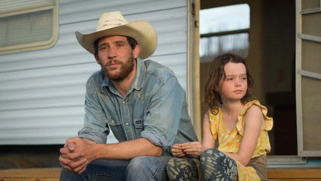 Man in cowboy hat and denim shirt sits beside young girl in yellow floral dress on steps of a trailer, both looking pensive.