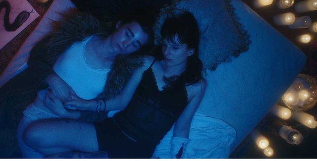 Two people lying close together on a bed bathed in blue light, surrounded by glowing candles, in an intimate nighttime scene.