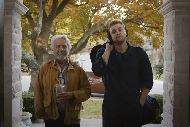 Two men on a stone porch: an older man with white hair in a tan jacket holding an iced drink, and a younger blond man with headphones around his neck carrying a backpack.