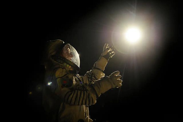 Astronaut in a spacesuit reaching toward a bright light source against a dark background, arms outstretched with gloved hands.