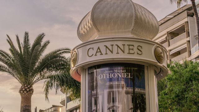 Ornate white kiosk with domed top labeled "CANNES" displaying a Jean-Michel Othoniel exhibition poster, with palm trees.