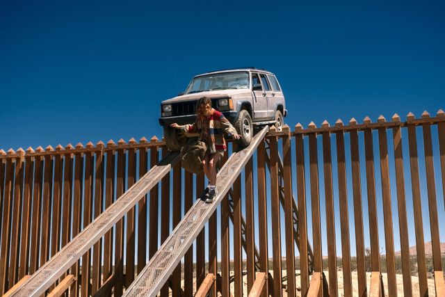 Person climbing a metal ramp over a tall brown border fence in a desert, with a Jeep Cherokee parked at the top.