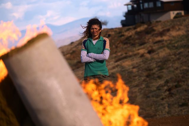 Woman with arms crossed wearing green vest and glasses stands on hillside behind burning debris, wind blowing her hair.