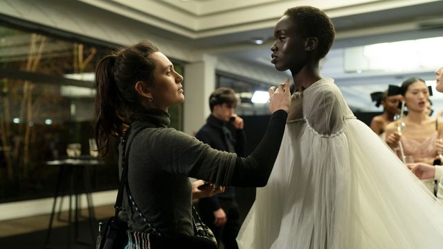 A couture fitting in progress, with a woman adjusting the bodice of a voluminous white dress on a model.