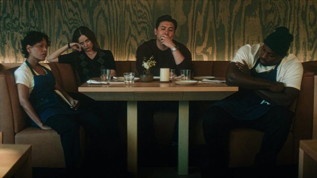 Four restaurant staff sit in a booth around a table, looking tired and contemplative in a dimly lit dining room.