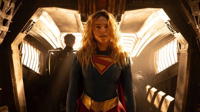 Supergirl stands in a warm-lit sci-fi interior wearing her blue suit with the red-and-gold S shield.