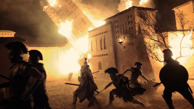 Silhouetted ancient warriors run through a burning city in a wide battle scene from The Odyssey.