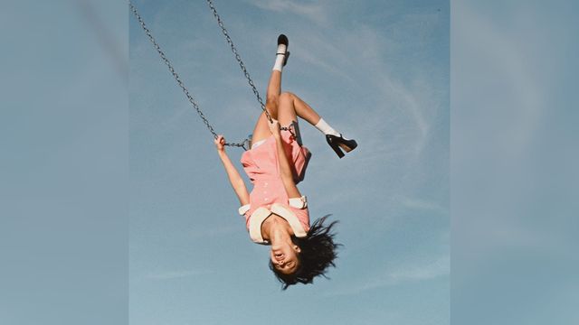 Olivia Rodrigo swings upside down in a pink outfit against a blue sky.