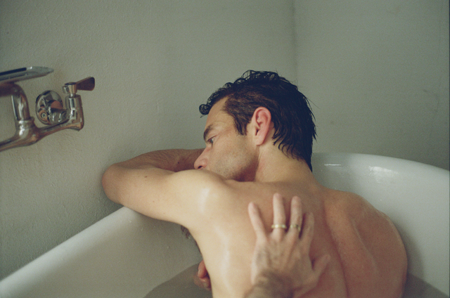 Shirtless man with wet hair sits in a bathtub beside another person’s hand resting on his back.