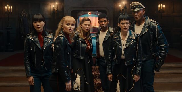Six people in studded black leather jackets stand tensely in a dim candlelit hall, with a glowing jukebox behind them.